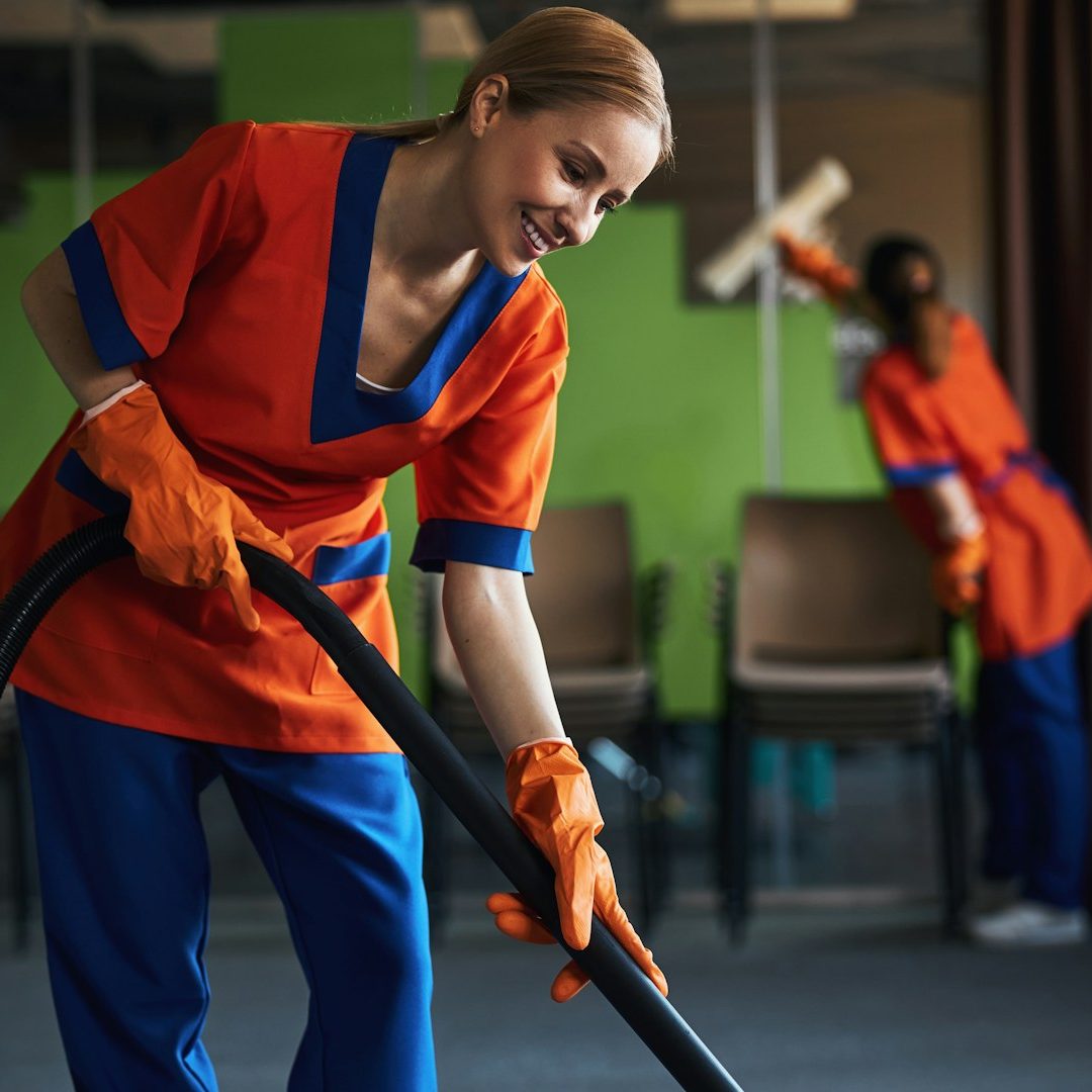 Deux femmes en uniforme nettoient une pièce, l'une passe l'aspirateur et l'autre frotte.